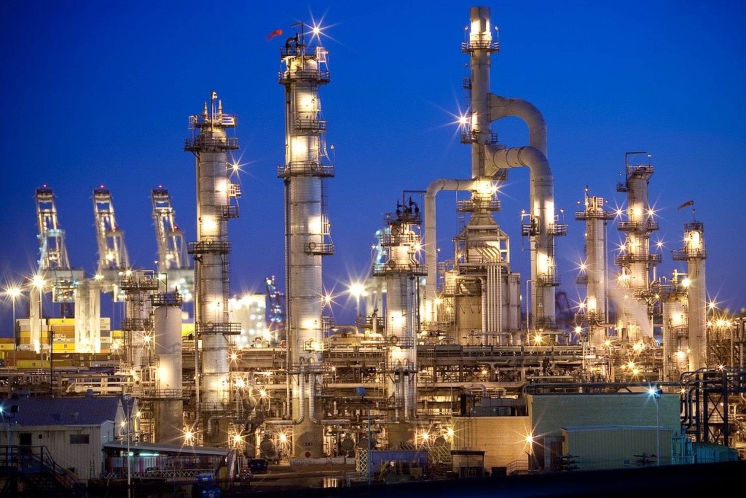 Industrial refinery with port cranes visible in the background.